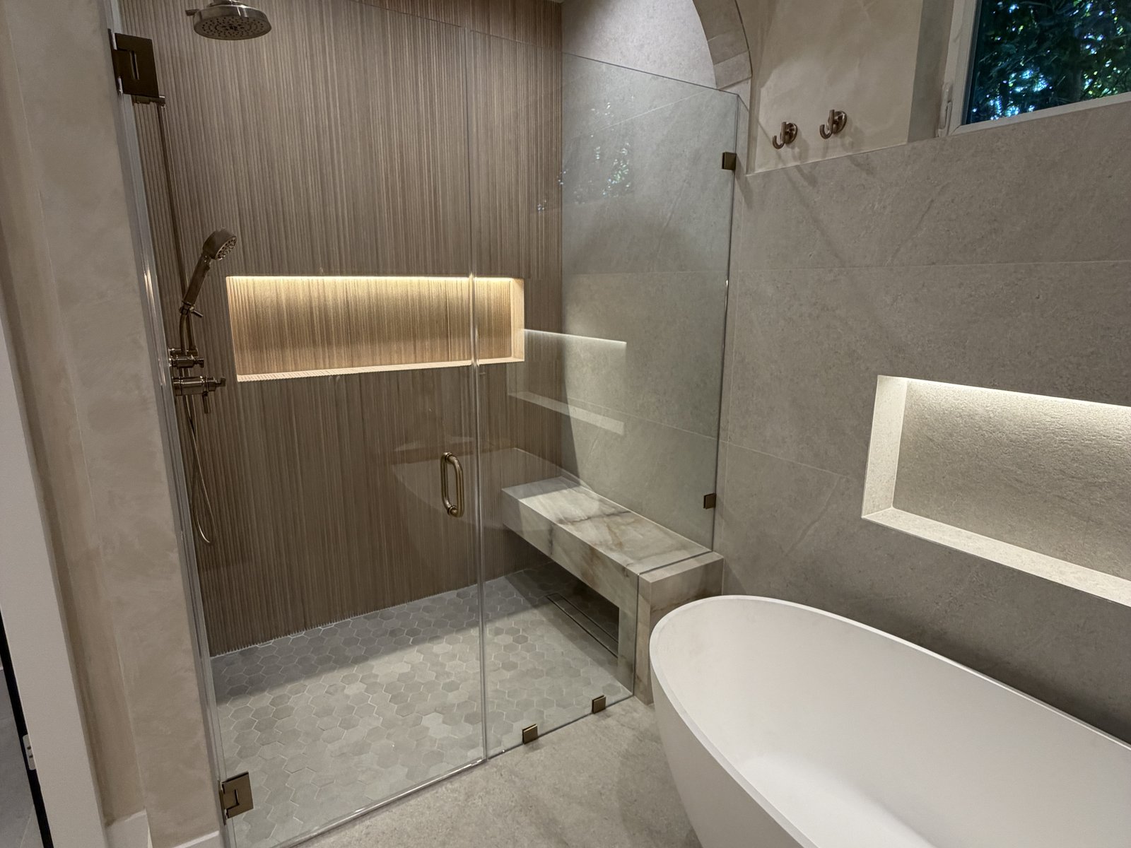 Finished bathroom with an arched tiled walk-in shower with a bench and lit niche behind a glass panel, next to a freestanding tub, completed by Home Pro Green Remodeling