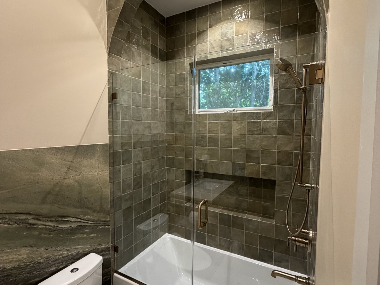 Finished bathroom with an arched tiled shower behind a glass door, a window above the shower and a wall-mounted toilet, completed by Home Pro Green Remodeling