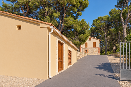 The restored estate at Mas El Regalo near Ibi: a cream-plastered outbuilding with a terracotta tiled roof, dark wooden door and white downspout beside a paved driveway climbing through tall pine trees to the two-storey stone main house with wooden shutters.