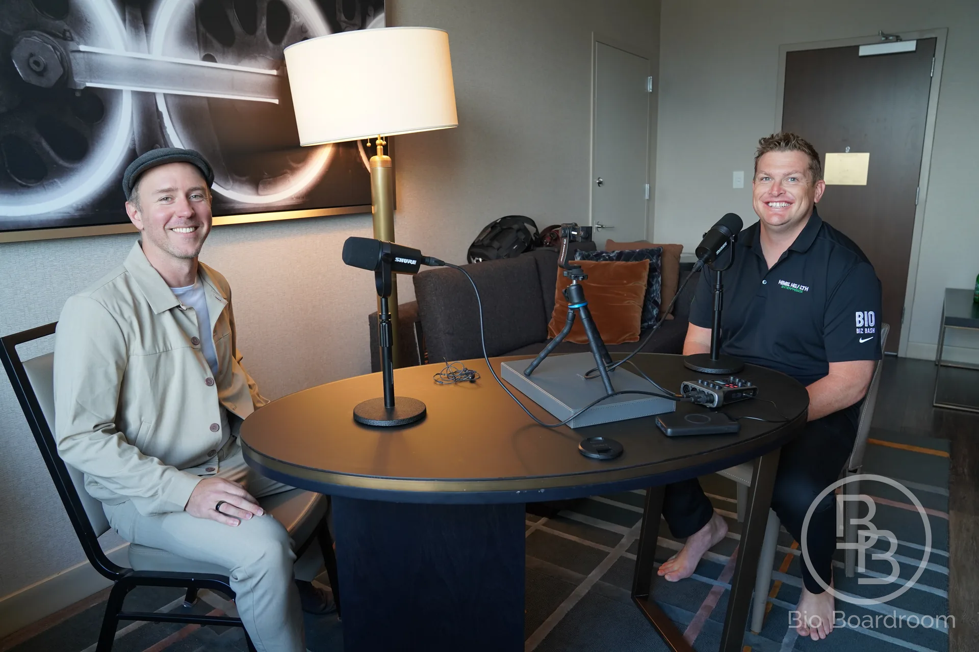 Two people seated at podcast microphones with a camera and audio recorder between them.