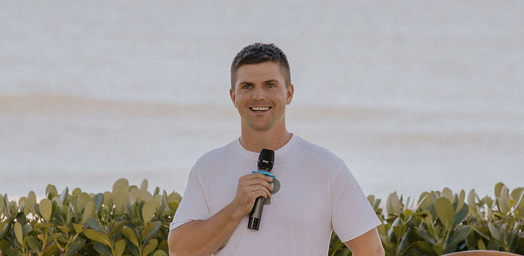 Jordan Dunin speaking to a group outdoors, handheld microphone in hand, a brass gong behind him