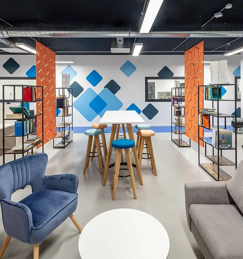 Contemporary open-plan office break-out area with a high table, colourful stools, soft seating and exposed services above