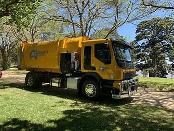 Yellow Wheelie Waste rear loader parked on grass with trees behind