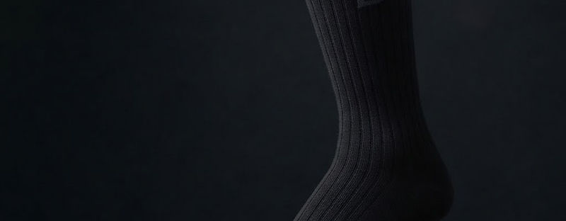 A single black ribbed crew sock with a tonal knitted B mark on the cuff, standing on a graphite slab, lit from the left with deep shadow behind