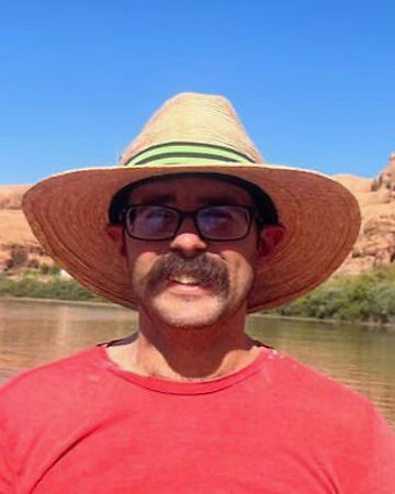 Ian Benedetti in a straw hat, glasses and red t-shirt with a cliff and water behind him
