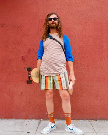 Ray Sumser standing against a terracotta wall in a striped raglan tee and sunglasses, holding a skateboard