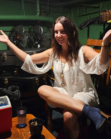 Kim Helmuth smiling in a white blouse, arms raised, sitting cross-legged on a wooden chair in a dark green studio