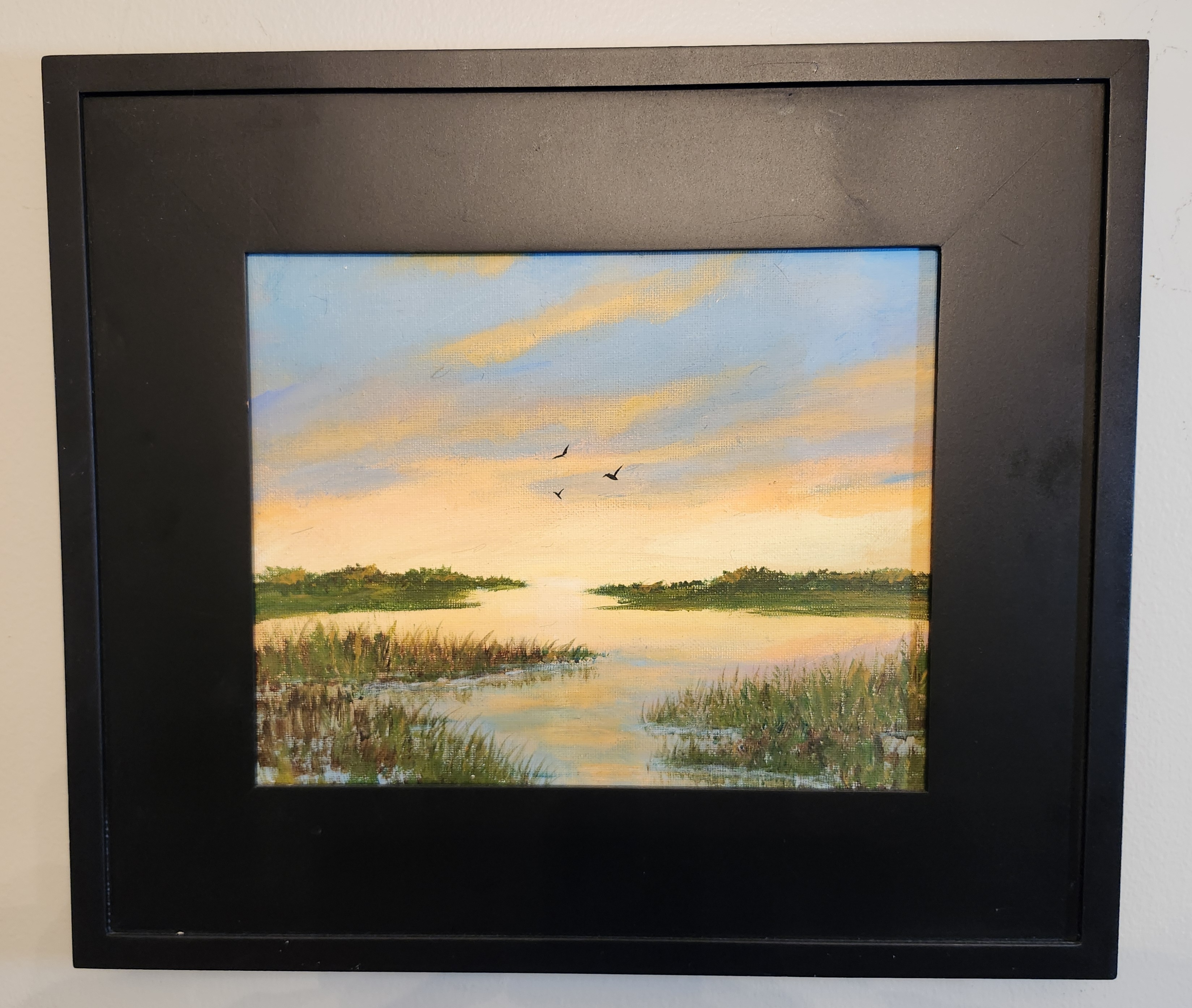 Marshland at sunset with a soft blue-to-peach sky, dark green tree line, reflective water and three bird silhouettes in the sky.