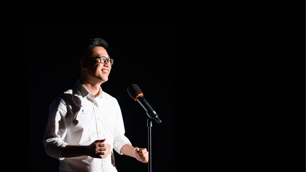Junhan Chin speaking on stage with a microphone, dramatically lit