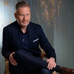 Portrait of Dan Skili, psychotherapist, seated and looking calmly toward the camera against a muted sage and gold textured wall.