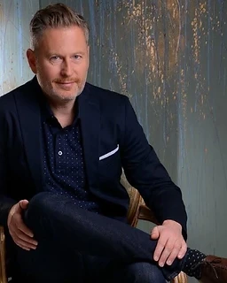 Portrait of Dan Skili, psychotherapist, seated and looking calmly toward the camera against a muted sage and gold textured wall.