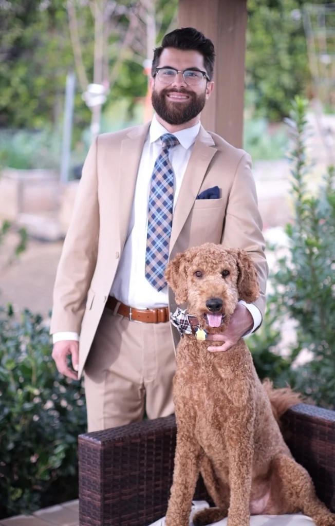 Devin Soliz, REALTOR® with Realty Masters & Associates, wearing a tan suit with his dog in an outdoor garden setting