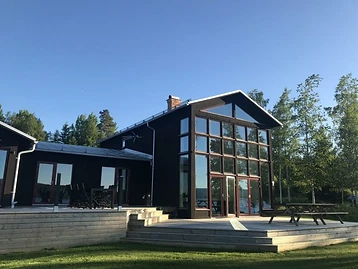Trähus med glasad gavelfasad och svart liggande panel omgivet av tallar.