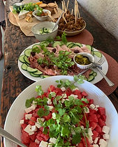 A long timber table set with a watermelon and feta salad, a platter of cold meats with cucumber and olives, and bowls of dips behind