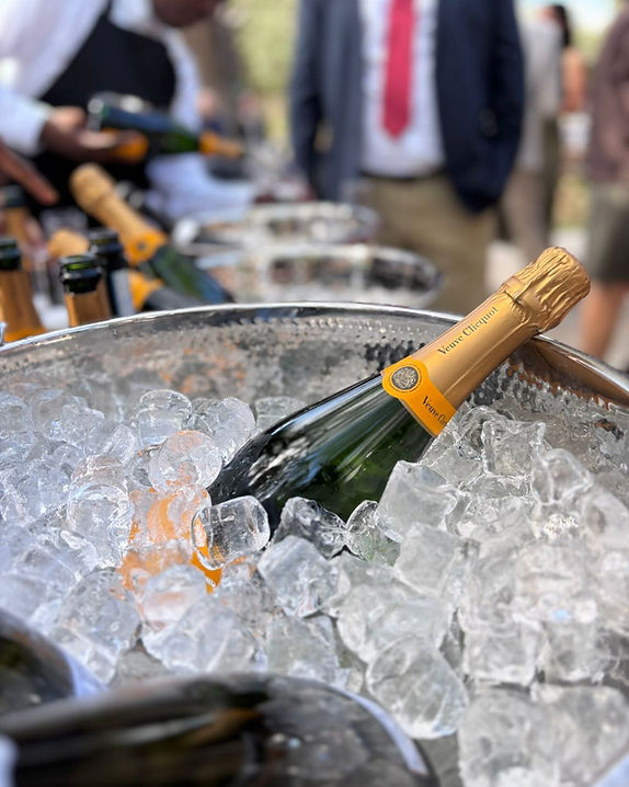 A bottle of champagne resting in a hammered metal ice bucket full of ice, with more bottles and guests out of focus behind