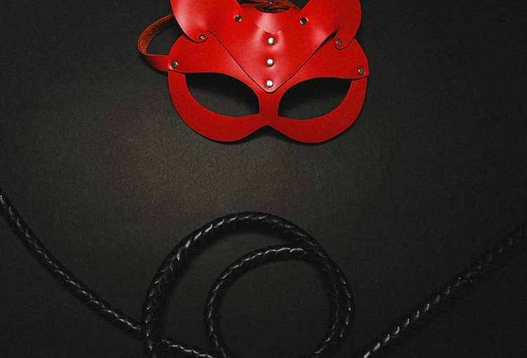 Red leather feline mask with silver rivets laid over a coiled black whip on charcoal