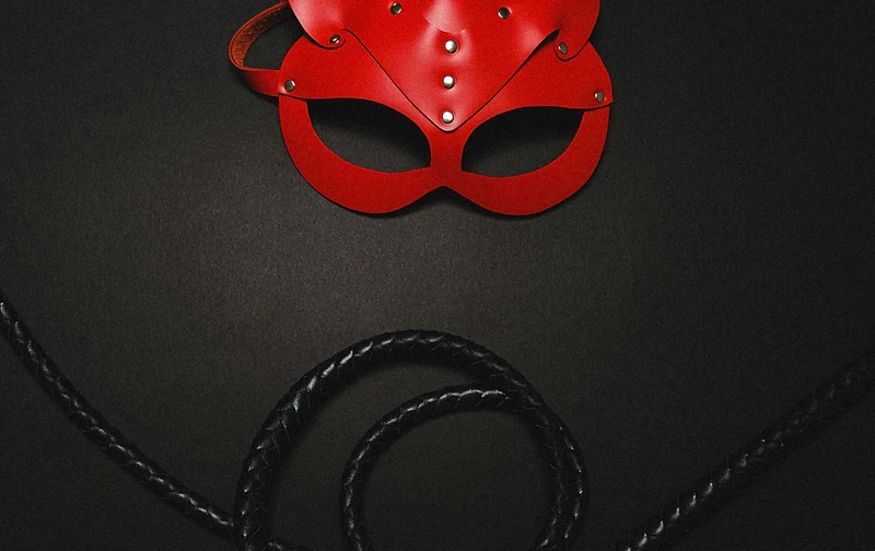 A red leather feline mask with silver rivets resting above a coiled black whip
