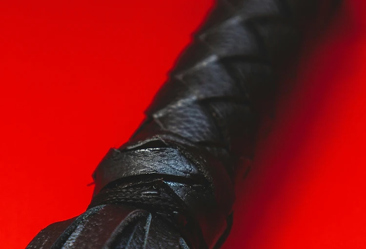 Braided black leather flogger handle against deep crimson — the mark of the house