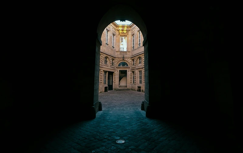 A dark stone archway opening onto a sunlit courtyard — the threshold before entry