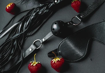 Flogger, ball gag, and blindfold arranged with red strawberries on matte black