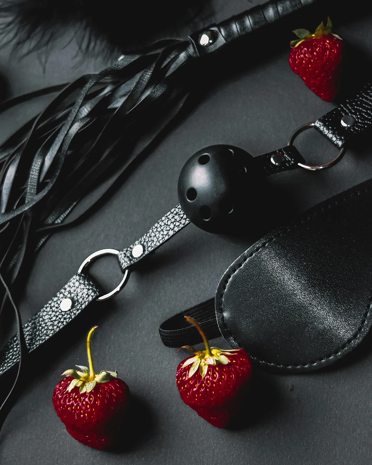 Flogger, ball gag, and blindfold arranged with red strawberries on matte black