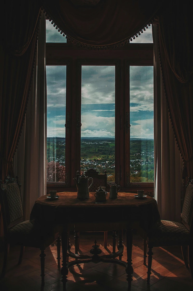 A table set for two in a shadowed salon, light falling through tall draped windows